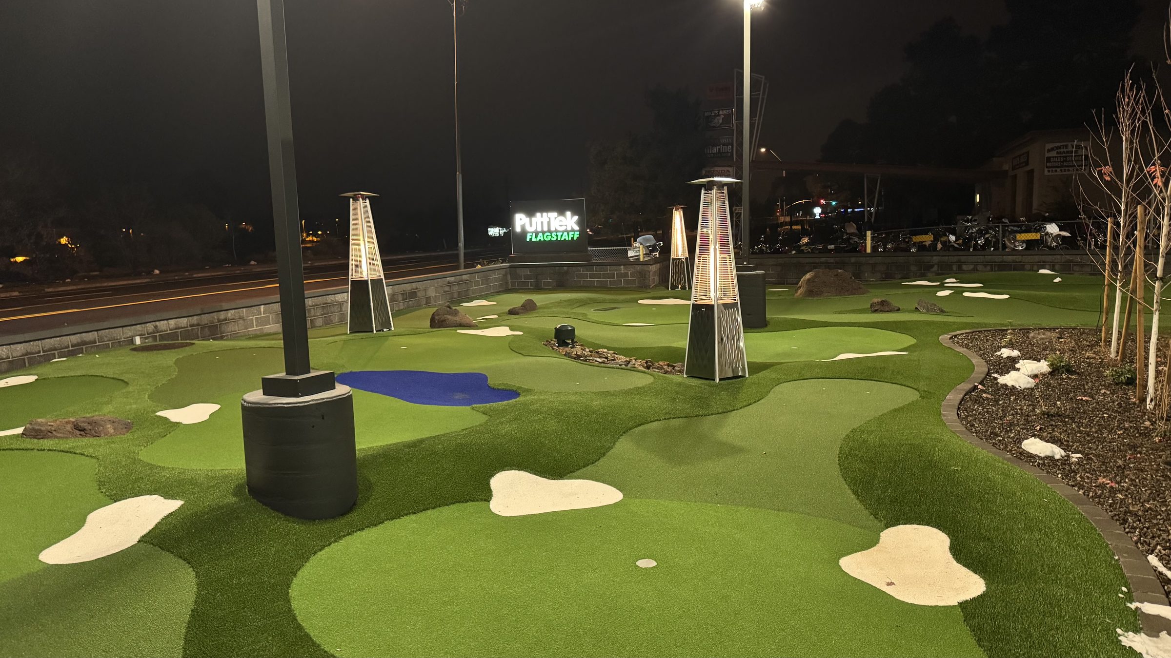 The PuttTek Flagstaff putting course lit up at night, with rolling contoured greens, sand-colored bunkers and patio heaters along Historic Route 66.