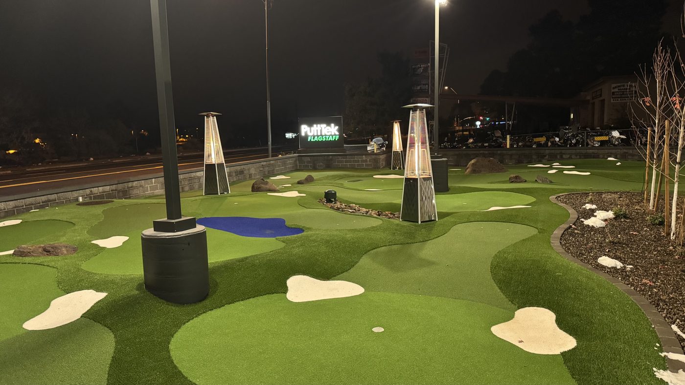 Wide view of the PuttTek Flagstaff greens at night, showing contoured turf, sand bunkers, a water feature and the lit PuttTek Flagstaff sign.